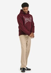 Kesten smeđa dukserica s velikim bijelim logotipom "TAPOUT", podesivim kapuljačom sa špagom, džepom na prednjoj strani, u kombinaciji sa bež hlačama i crnim tenisicama.