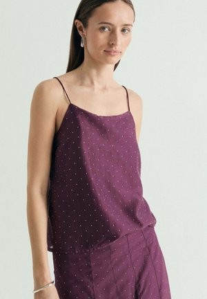 Mujer con top morado sin mangas y pantalones a juego con pequeños lunares blancos, de pie frente a un fondo claro y liso.