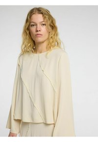 Lys cremefarvet top med lange ærmer, der har en løs pasform, slidte sømme og en rund halsudskæring. Letvægts, glat stof med minimal detaljer.