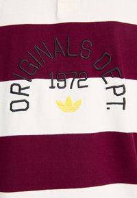 Bordó és fehér csíkos póló ing fekete hímzett "ORIGINALS DEPT. 1972" felirattal és alatta egy sárga Adidas trefoil logóval.
