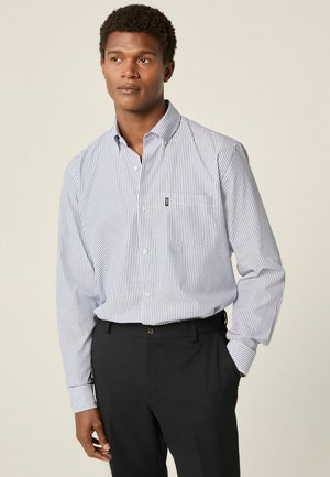 Chemise à rayures bleu et blanche avec un col, manches longues et poche poitrine, associée à un pantalon sombre bien ajusté.