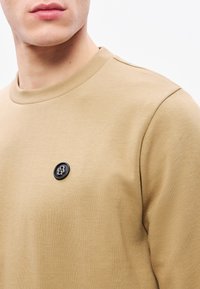 Béžová mikina s okrúhlym výstrihom, na hrudi je čierne kruhové logo, vyrobená z textúrovaného bavlneného zmesi. Minimalistický dizajn.