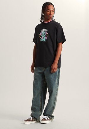 T-shirt grafica nera con design di un personaggio dei cartoni animati colorato, abbinata a jeans larghi azzurri e sneakers con motivo.