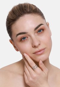Anello d'argento con filamenti intrecciati, caratterizzato da una finitura lucida. Il modello si tocca il viso, mettendo in evidenza l'anello. Sfondo neutro.
