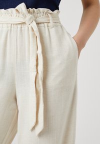Pantalon en lin beige avec une taille froncée et une ceinture nouée. Doté de poches latérales et d'un tissu texturé, mettant en valeur un design décontracté.