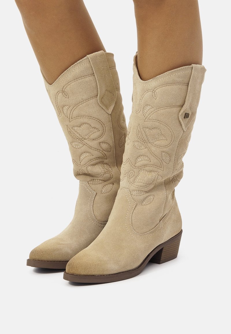 Bottes de cowboy en suède beige avec un bout pointu, des coutures western et un petit talon carré. Accentuation avec des motifs floraux en relief.