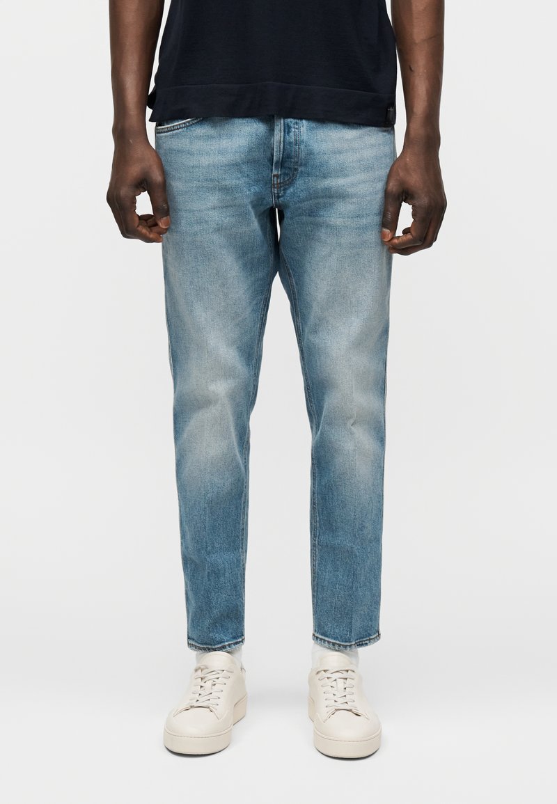 Homme portant un jean délavé bleu clair, des baskets blanches et un t-shirt noir à manches courtes, debout devant un fond clair uni.
