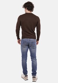 Uomo con capelli ricci che indossa una camicia a maniche lunghe marrone, jeans blu e sneaker bianche, in piedi di spalle su uno sfondo neutro.