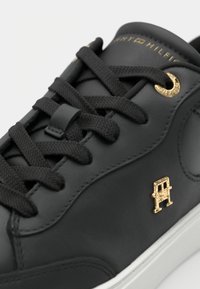 Fekete bőr sportcipő arany részletekkel, fekete fűzőkkel, kerek fűzőlyukakkal és fehér talppal. Jellegzetes logórészlet az oldalon.