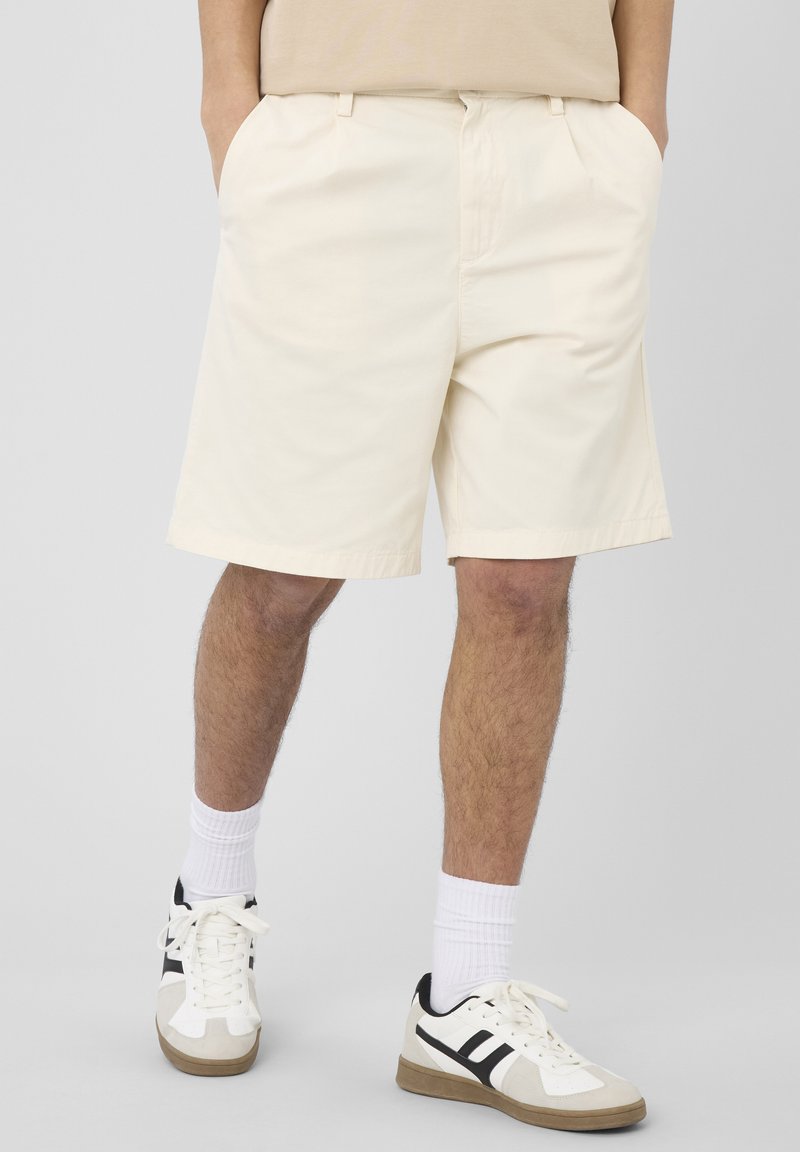 Homme portant un short beige, des chaussettes blanches et des baskets blanches avec des accents noirs, debout les mains dans les poches.