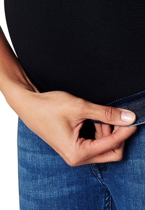 Dark blue denim jeans with a stretch band, featuring a smooth texture. A hand grips the waistband, showing the fabric's flexibility.