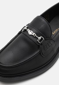 Mocassino in pelle nera perforata con un accento in catena argentata e una texture liscia. Presenta una punta arrotondata e dettagli cuciti lungo i lati.