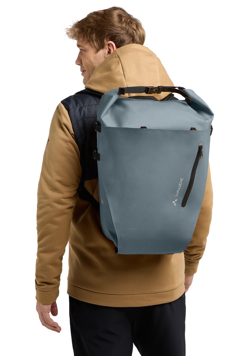 Lichtblauwe waterdichte rugzak met een oprolsluiting, zijvak met rits en verstelbare schouderbanden, gedragen door een persoon in een tan-kleurige sweatshirt.