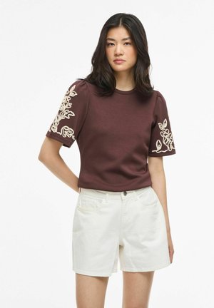 Giovane donna con capelli scuri che indossa una maglietta marrone a maniche corte con ricamo floreale bianco sulle maniche e pantaloncini bianchi a vita alta.