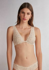 Beige kanten bralette met een diepe V-halslijn, golvende randen, bloemenpatronen en een centrale strikdetail. Sluiting aan de achterkant.