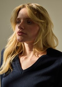 Pull en maille bleu marine foncé à col en V, fabriqué en tissu doux, avec des détails côtelés au niveau du col. De longs cheveux tombent sur une épaule. Un éclairage naturel met en valeur la texture.