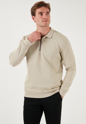 Beiger Pullover mit einem Reißverschlusskragen, langen Ärmeln und gerippten Bündchen. Hergestellt aus einem glatten, strukturierten Stoff, kombiniert mit schwarzen Hosen.