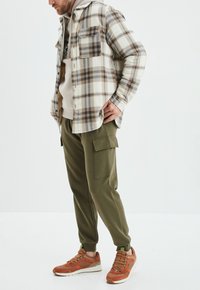 Camicia di flanella a quadri beige con due tasche sul petto, indossata sopra un felpa crema con cappuccio. Pantaloni cargo verde oliva con tasche laterali e sneaker arancioni.