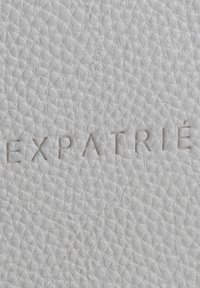 Surface en cuir blanc texturé avec le mot "EXPATRIÉ" embossé en lettres majuscules, centré près du bas.