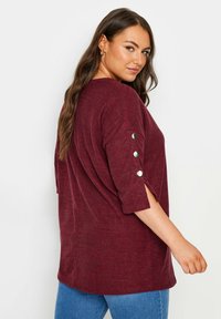 Burgundy ykköslangan ylisuuri paita, jossa on puolihihat ja napitakeet hihoissa. Pehmeä, teksturoitu kangas, jossa on sivukerat. Yhdistetty sinisiin farkkuihin.