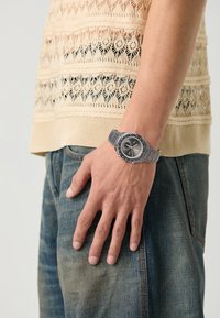 Montre en métal argenté avec un cadran noir rond, des index contrastants et un bracelet en caoutchouc gris, portée sur une main contre un fond clair.