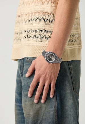 Montre en métal argenté avec un cadran noir rond, des index contrastants et un bracelet en caoutchouc gris, portée sur une main contre un fond clair.
