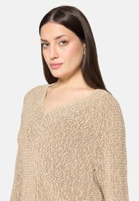 Pull en maille beige avec un col en V, un motif texturé et une coupe ample. Le tissu semble doux et léger. Couture détaillée sur l'ensemble.