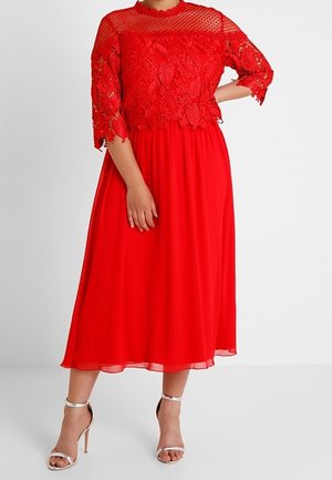 Kobieta w czerwonej sukience midi z koronkowym kwiatowym topem i przezroczystymi rękawami do łokcia, w połączeniu z srebrnymi sandałami na wysokim obcasie.