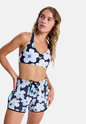 Frau trägt marineblaue Sport-BH mit Blumendesign und passende Shorts mit Kordel, posiert mit einer Hand auf der Hüfte vor weißem Hintergrund.