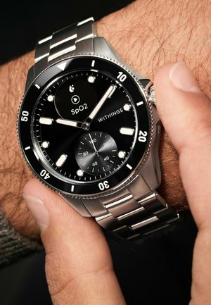 Nahaufnahme eines Handgelenks mit einer Edelstahl-Withings-Uhr, die SpO2-Gesundheitsdaten anzeigt, während Hände die Krone der Uhr einstellen.