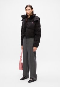 Casaco curto acolchoado preto com fecho de zip e botões de pressão, combinado com calças cinzas listradas de cintura alta e uma bolsa de ombro rosa. Sapatilhas pretas.