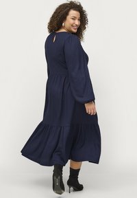 Vestido azul marino de mangas largas con un dobladillo en capas, tejido texturizado y detalle en forma de ojo de cerradura en la parte posterior. Se lleva con botines negros de tacón.