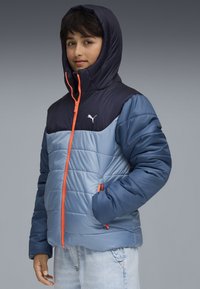 Blaues und marinefarbenes Steppjacke mit Kapuze, verziert mit orangen Reißverschlüssen und einem Logo, gesteppter Textur und seitlichen Taschen.