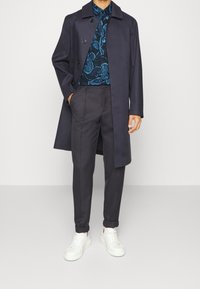 Marineblauwe jas met dubbele knopen, blauw shirt met bloemen, donkere broek en witte sneakers, wat een gelaagd, contrasterend outfit laat zien.