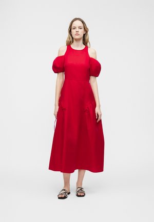 Kvinde iført en rød cold-shoulder midikjole med puffede ærmer og sorte og hvide slip-in sandaler med slangeprint, stående foran en ensartet grå baggrund.