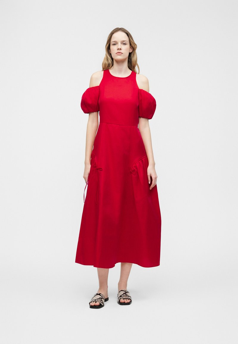 Γυναίκα που φοράει midi φόρεμα κόκκινου χρώματος με ανοιχτούς ώμους και φουσκωτά μανίκια, καθώς και δερμάτινα σανδάλια με ασπρόμαυρο σχέδιο φίδι, στέκεται σε απλό γκρι φόντο.