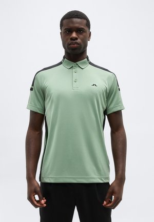 Junger Mann trägt ein hellgrünes Polohemd mit schwarzen Akzenten, steht mit entspannten Armen vor einem einfarbigen grauen Hintergrund.