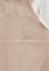 Rosa Corduroy-Latzhose mit vertikaler Textur; auf der Vorderseite ist ein gesticktes Hunde-Gesicht zu sehen. Ein weißes Shirt blitzt von der Seite hervor.