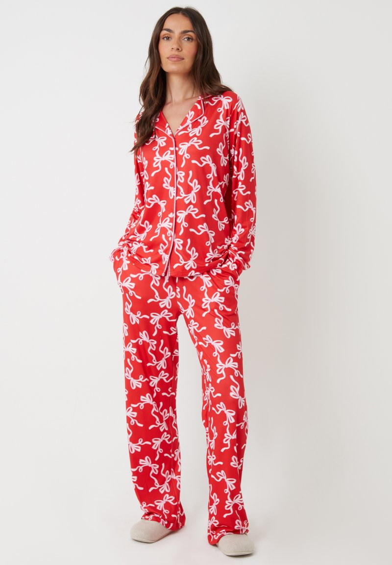 Rode pyjama-set met een wit strikpatroon, lange mouwen en een knoopsluiting shirt met zakken, gecombineerd met bijpassende broek en grijze pantoffels.