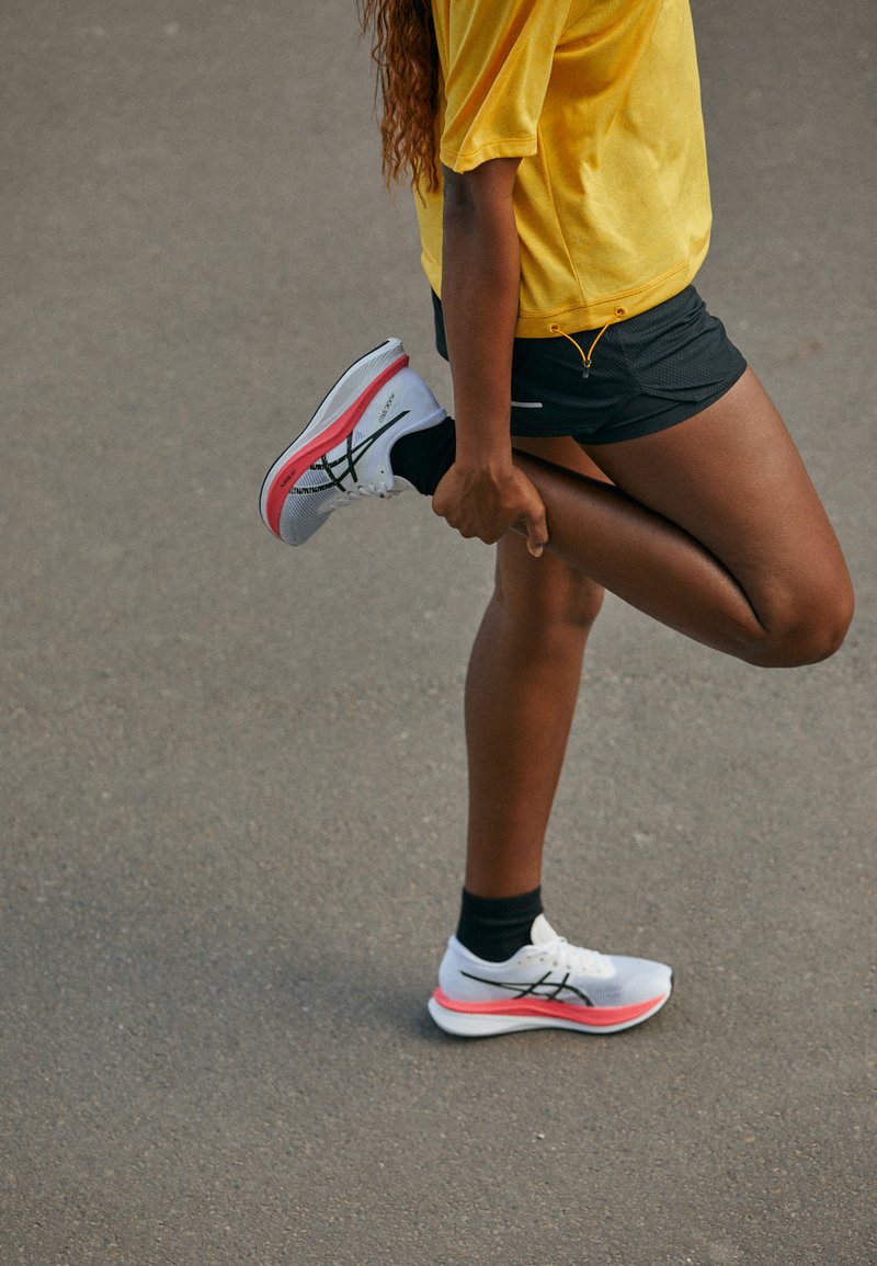 Baskets de sport grises avec des accents noirs et des semelles roses, portées avec des chaussettes noires, accompagnées d'un t-shirt jaune et d'un short noir.