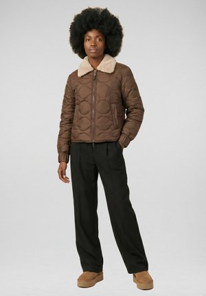 Femme portant une veste matelassée marron avec un col en fausse fourrure beige, un pantalon large noir et des chaussures compensées beige, debout devant un fond uni.