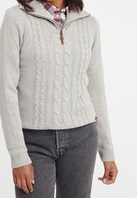 Pullover grigio chiaro in maglia con motivo a treccia, colletto con zip e dettagli in legno. Abbinato a jeans grigi scuri che evidenziano una vestibilità aderente.