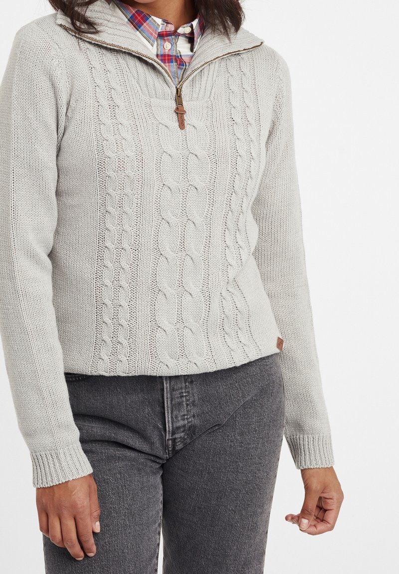 Pullover grigio chiaro in maglia con motivo a treccia, colletto con zip e dettagli in legno. Abbinato a jeans grigi scuri che evidenziano una vestibilità aderente.