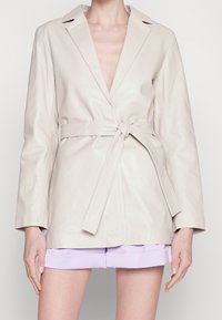 Veste enveloppante en cuir beige avec manches longues, décolleté plongeant en V et taille nouée. Texture lisse et coupe ajustée avec revers crantés.