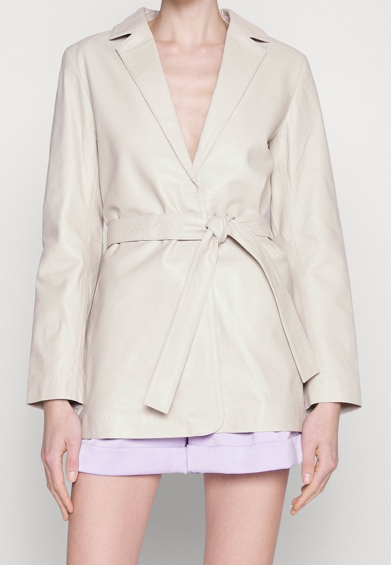 Veste enveloppante en cuir beige avec manches longues, décolleté plongeant en V et taille nouée. Texture lisse et coupe ajustée avec revers crantés.