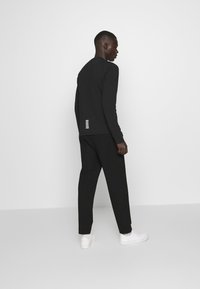 Maglietta nera a maniche lunghe con strisce orizzontali bianche sul retro, abbinata a pantaloni neri e sneakers bianche. Design semplice, vestibilità slim.