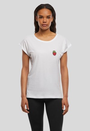 Femme portant un t-shirt blanc avec un petit motif de fraise rouge sur la poitrine, associé à un legging noir, debout sur un fond gris.