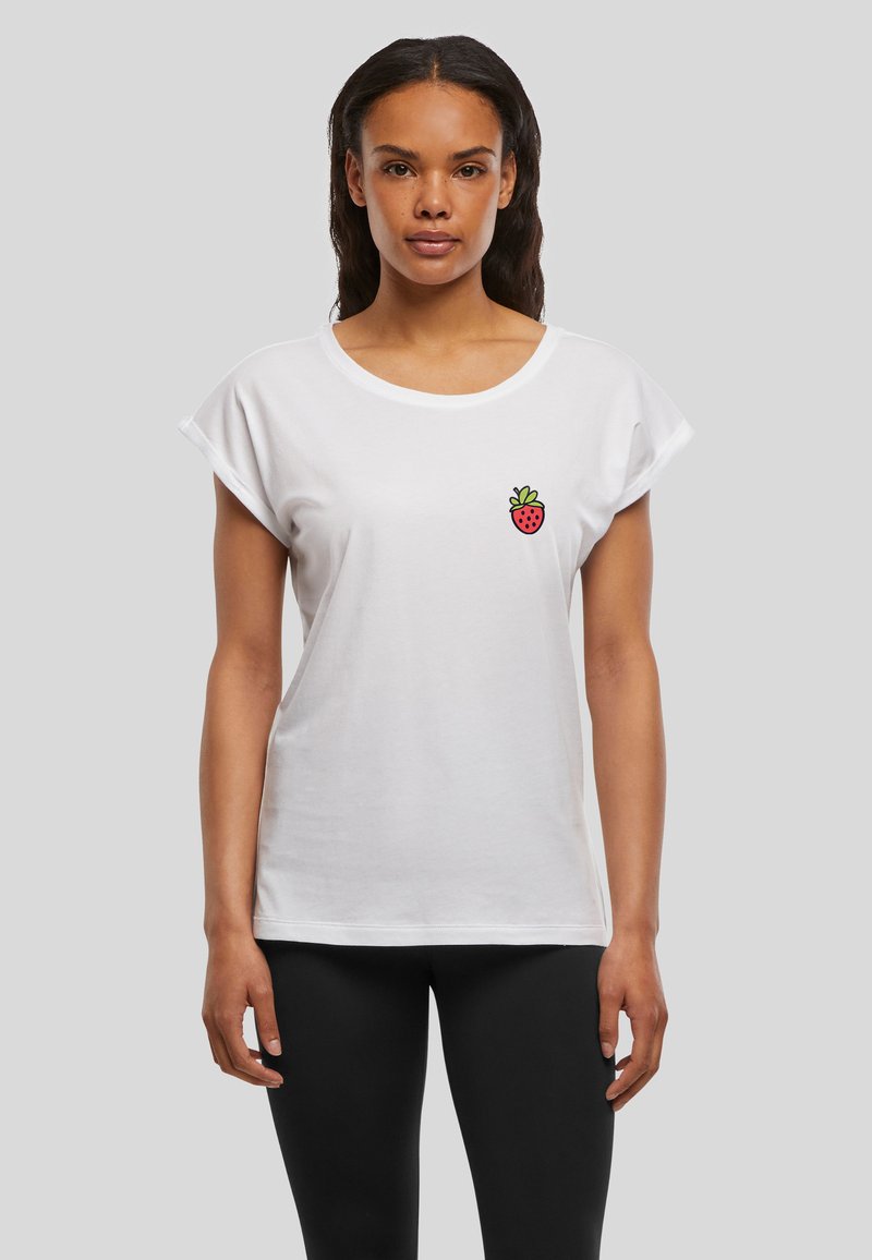 Femme portant un t-shirt blanc avec un petit motif de fraise rouge sur la poitrine, associé à un legging noir, debout sur un fond gris.