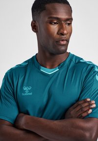 Turkis atletisk t-shirt med korte ærmer, rund hals og kontrasterende skulderdetaljer. Har et diskret logo på brystet.