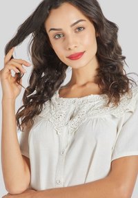 Blusa de crema ligera con mangas cortas, cierre de botones y delicados detalles de encaje en el escote. Textura suave, diseño casual.